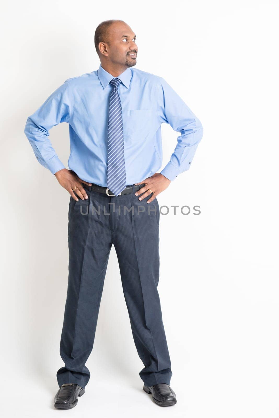 Portrait of full body mature Indian business man hands on waist looking to side, standing on plain background.