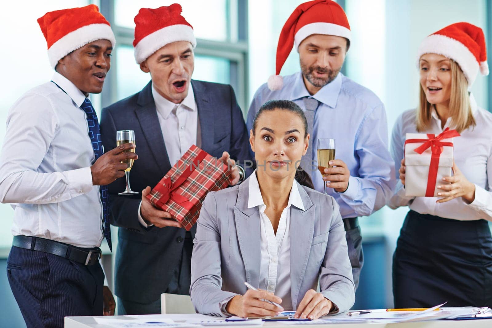 Amazed businesswoman looking at camera at workplace while colleagues with gifts and champagne congratulating her behind