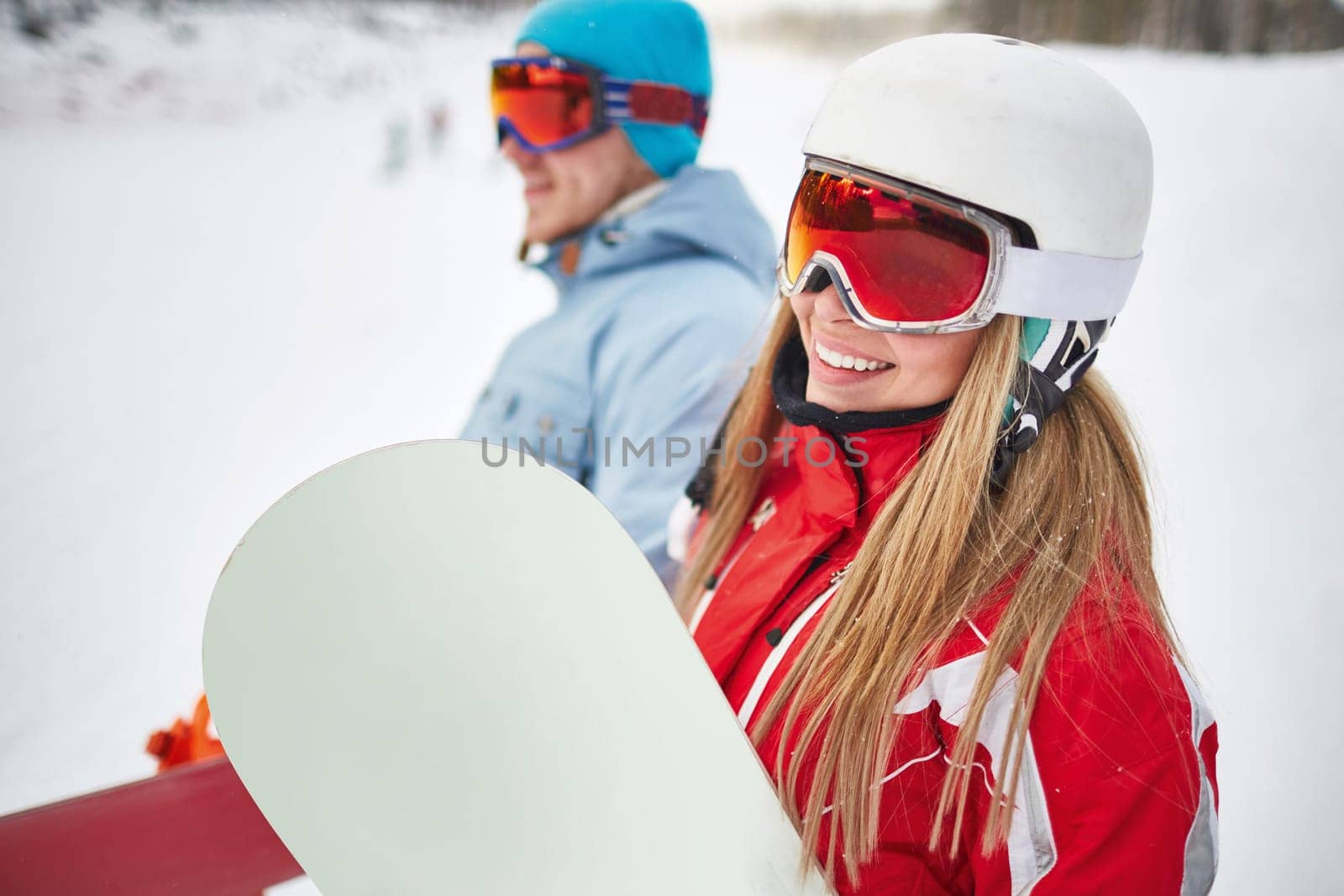Happy young snowboarder in activewear looking at camera on background of her boyfriend on winter resort