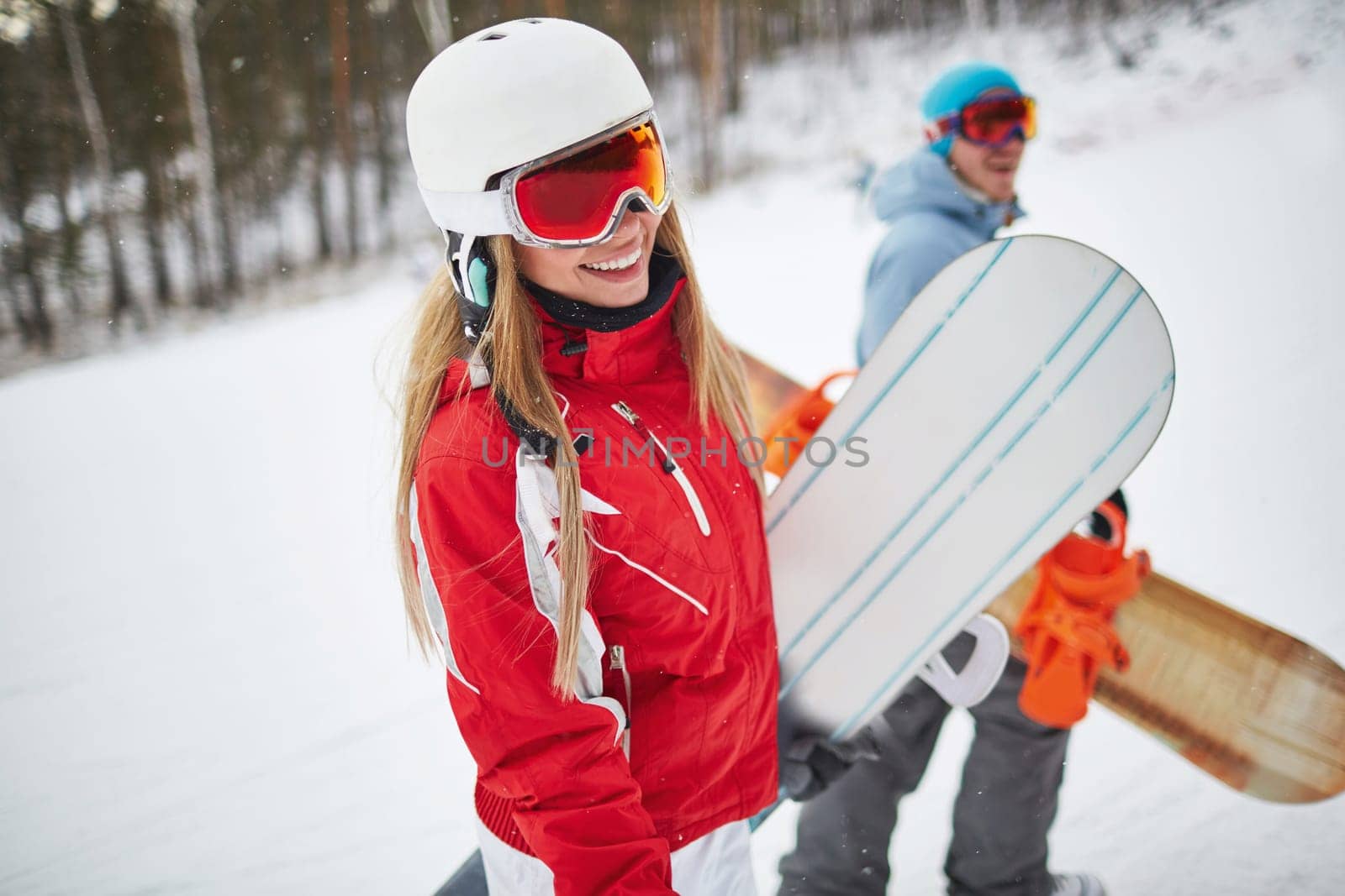 Young female with snowboard looking at camera on background of her boyfriend on winter resort