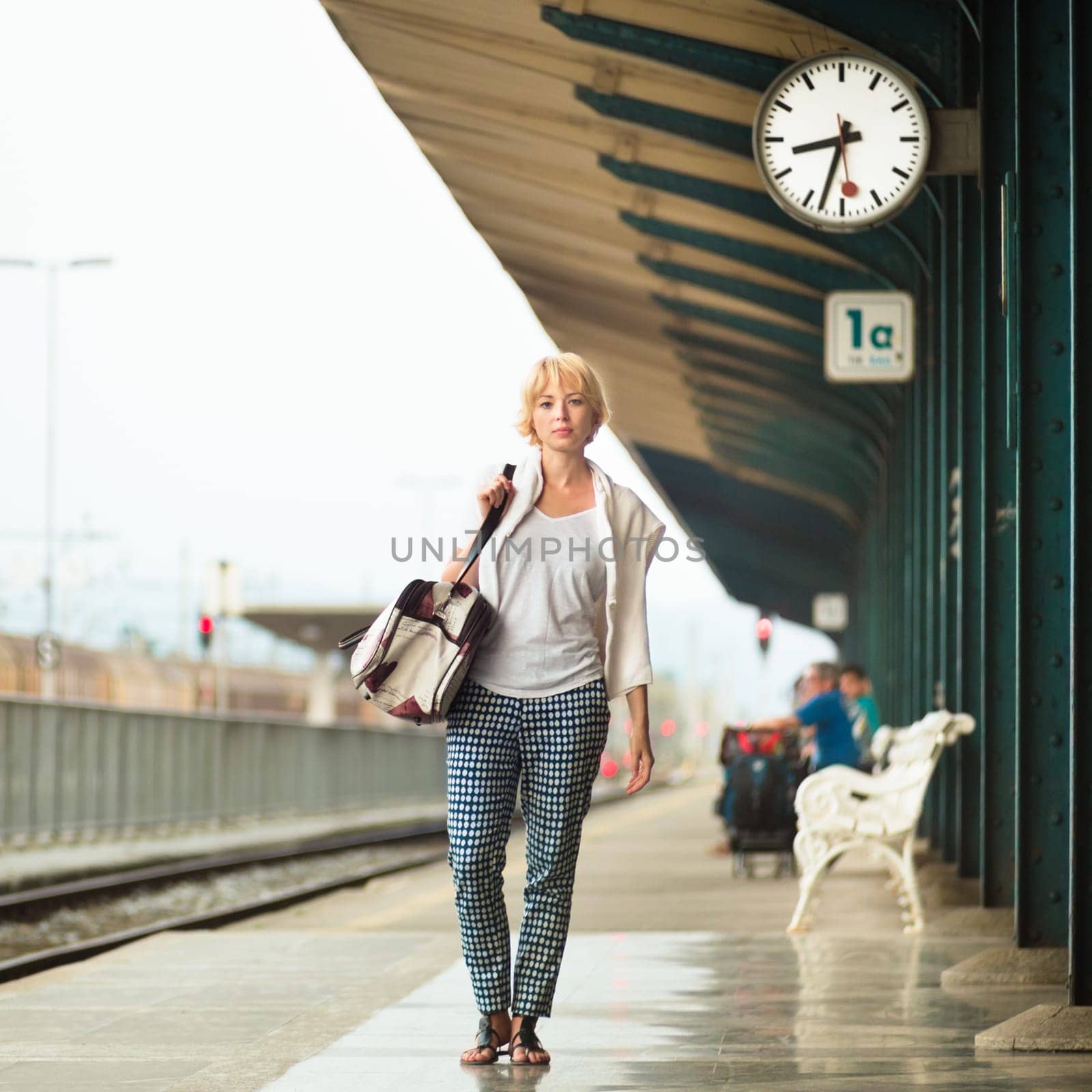 Blonde caucasian woman waiting at platform of railway station bearing vintage shoulder bag. Square composition.
