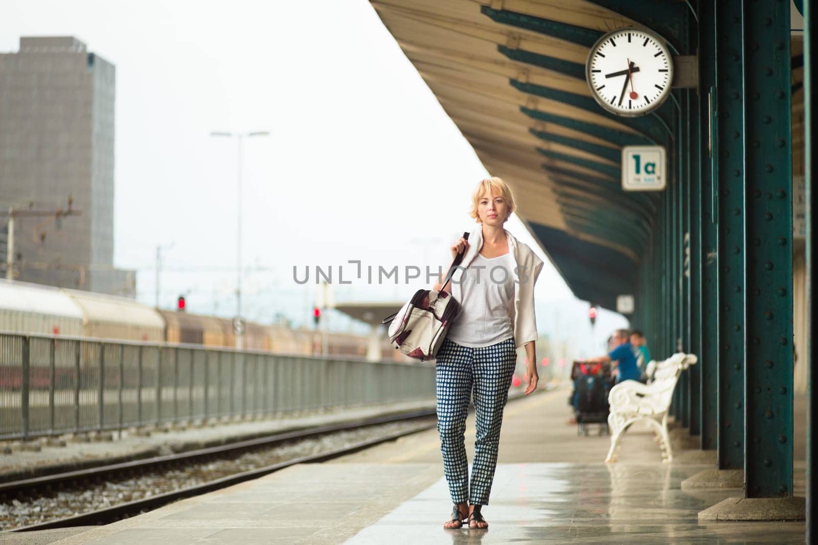 Blonde caucasian woman waiting at platform of railway station bearing vintage shoulder bag.