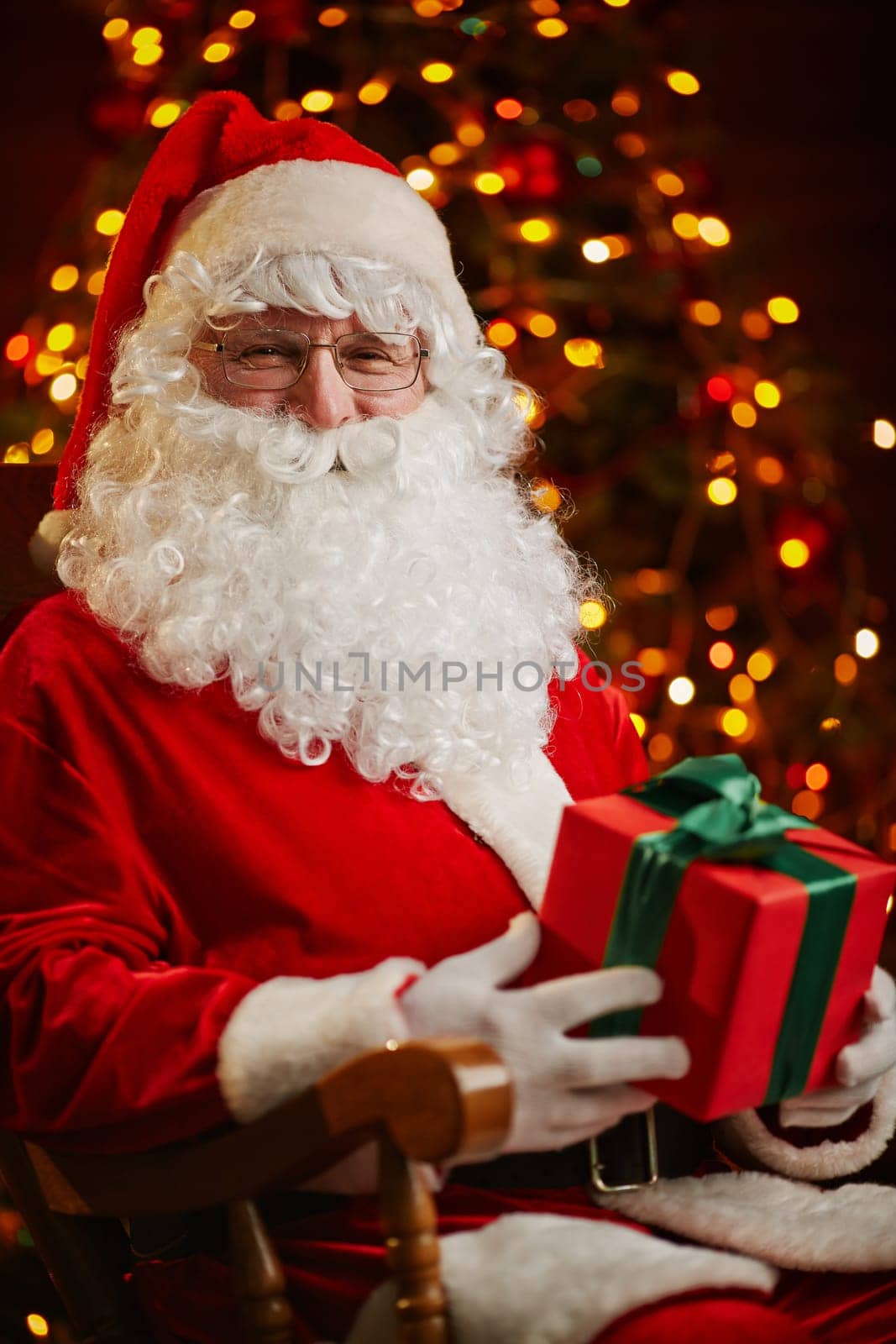 Happy Santa Claus with package looking at camera on background of sparkling firtree