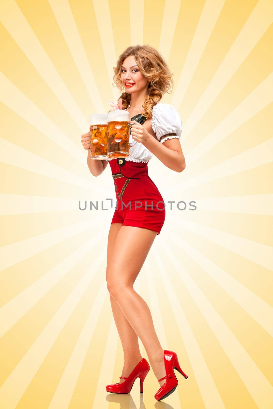 Beautiful tempting sexy woman wearing red jumper shorts with suspenders as traditional dirndl, serving two beer mugs with smile on colorful abstract cartoon style background.
