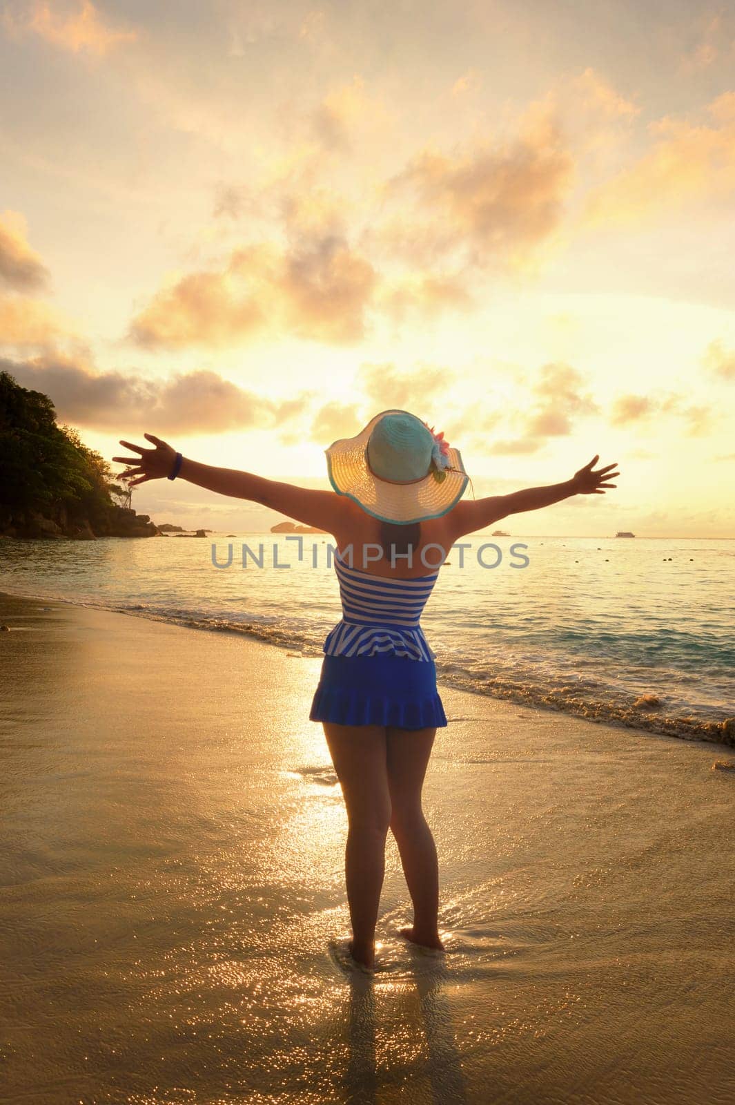 Tourist girl in a swimsuit blue and white striped hat standing on beach with pleasure the beautiful landscape of sky and sea during sunrise, Miang island, Similan National Park, Phang Nga, Thailand