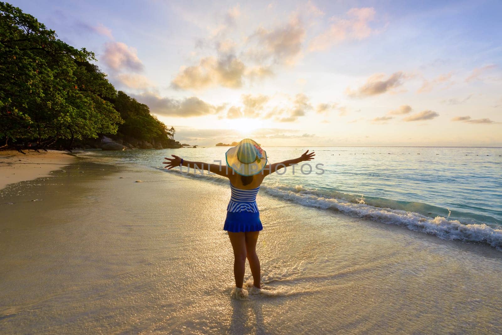 Tourist girl in a swimsuit blue and white striped hat standing on beach with pleasure the beautiful landscape of sky and sea during sunrise, Miang island, Similan National Park, Phang Nga, Thailand