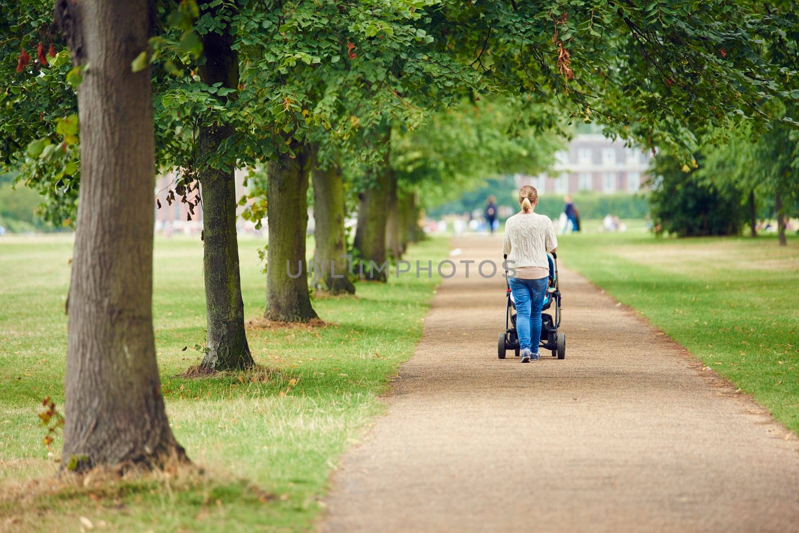Unrecognisable woman pushing pram walking down tree-lined path in park.