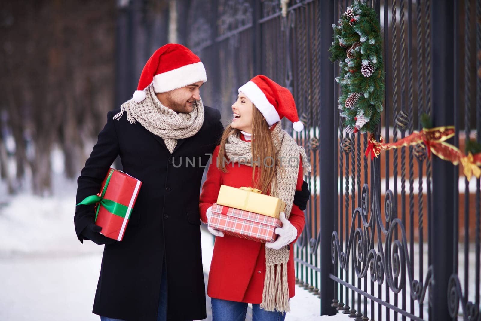 Affectionate couple in Santa caps and coats coming back home with presents for Christmas