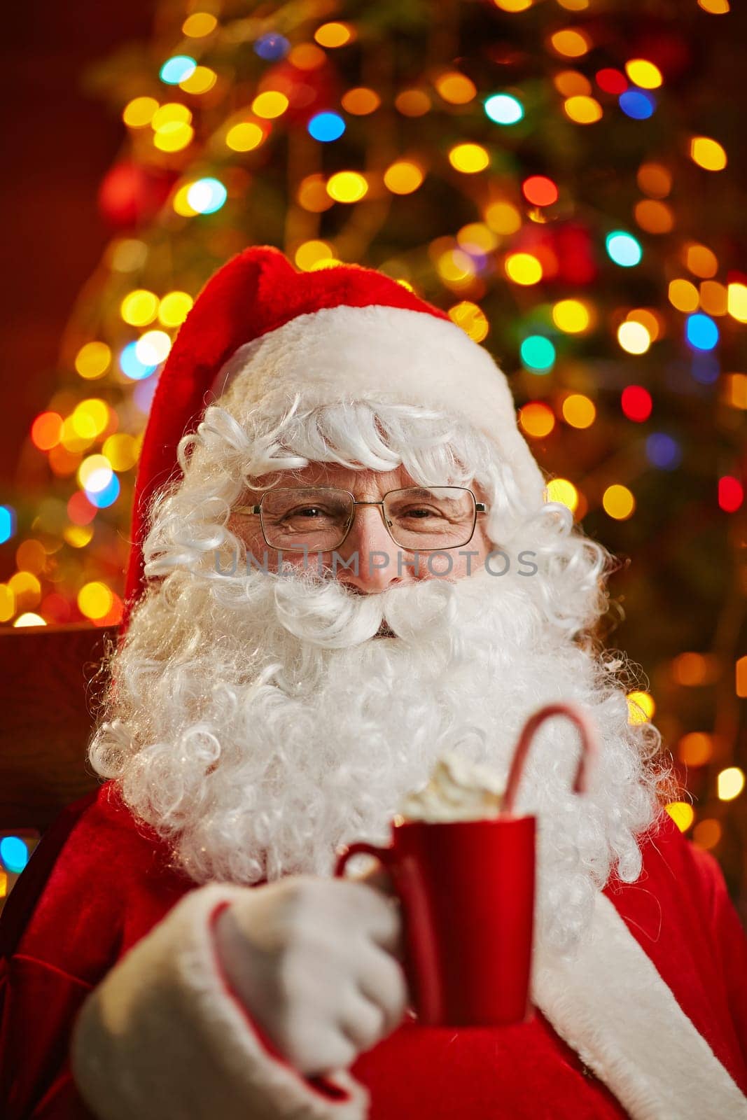 Santa Claus holding red cup with latte on background of sparkling xmas tree
