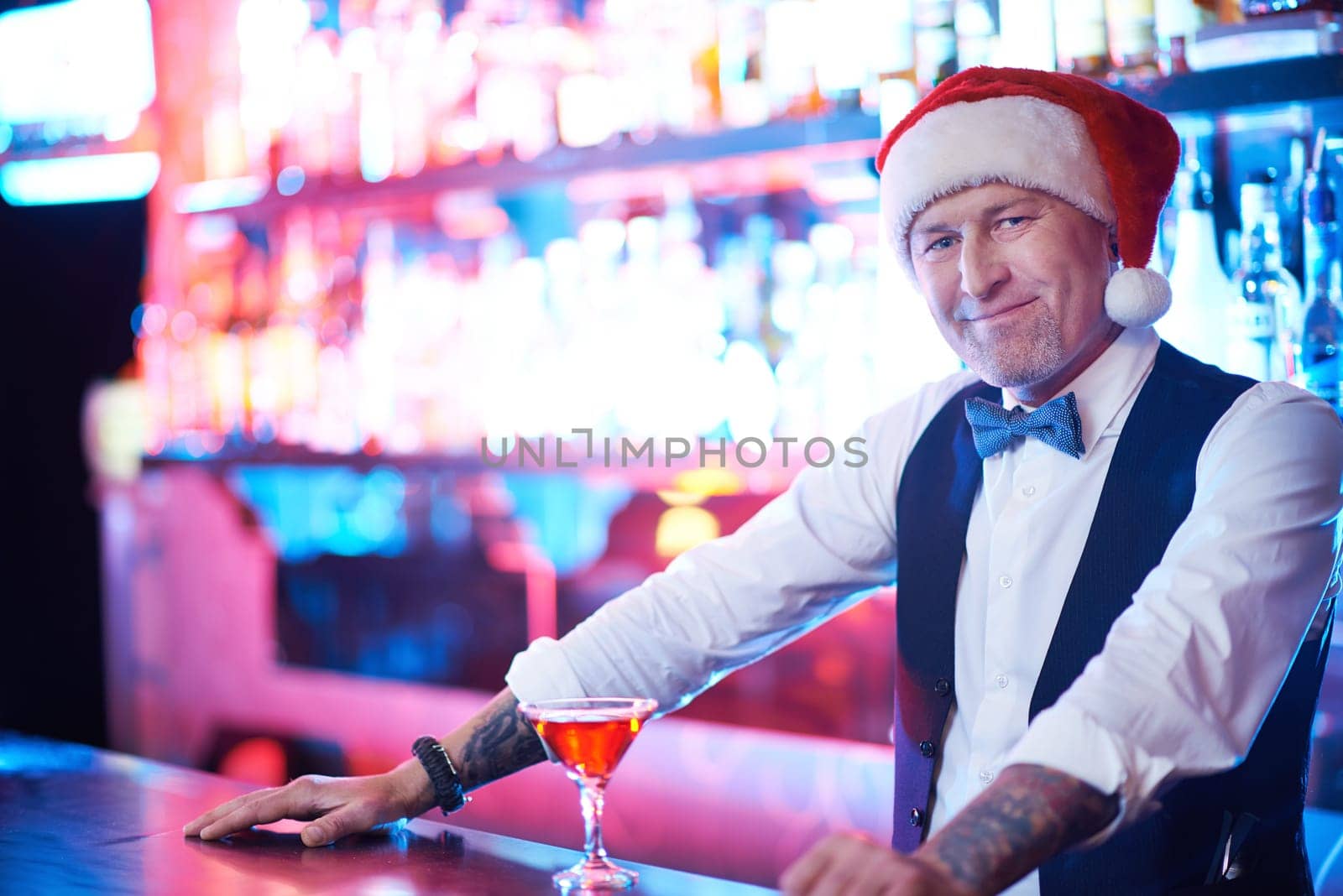 Happy barman in Santa cap standing by counter with martini and looking at camera