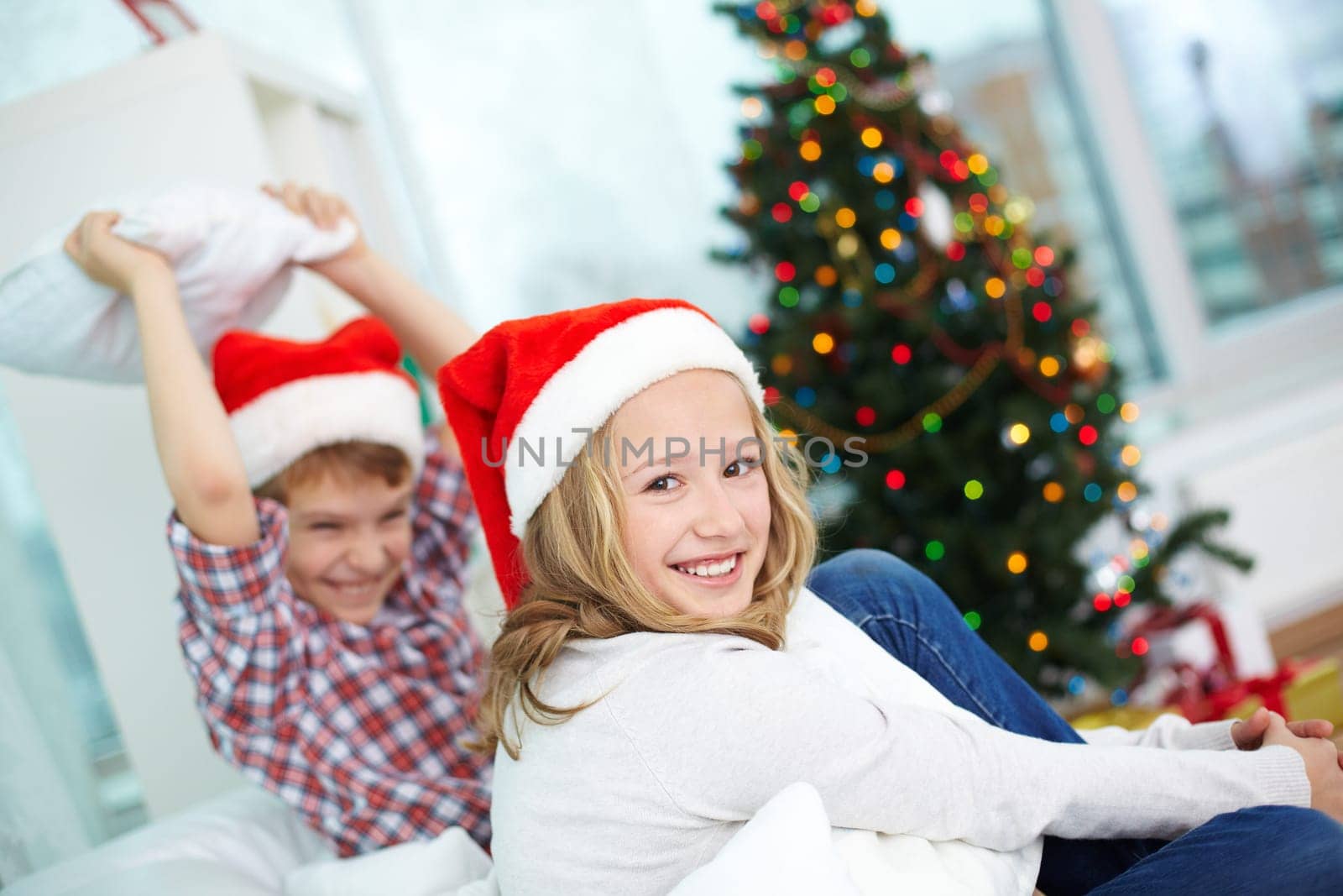 Happy girl in Santa cap looking at camera on background of her brother on Christmas evening