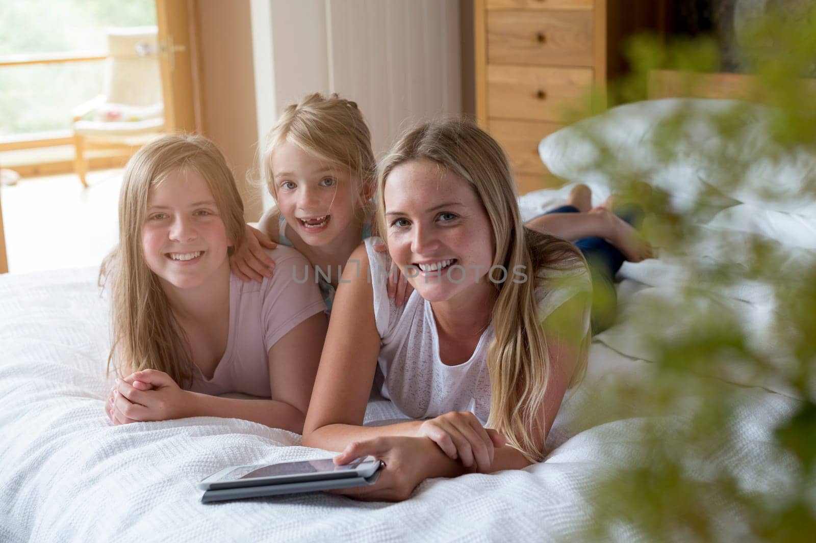 Sisters lying on a bed in their home with a digital tablet, smiling for the camera.