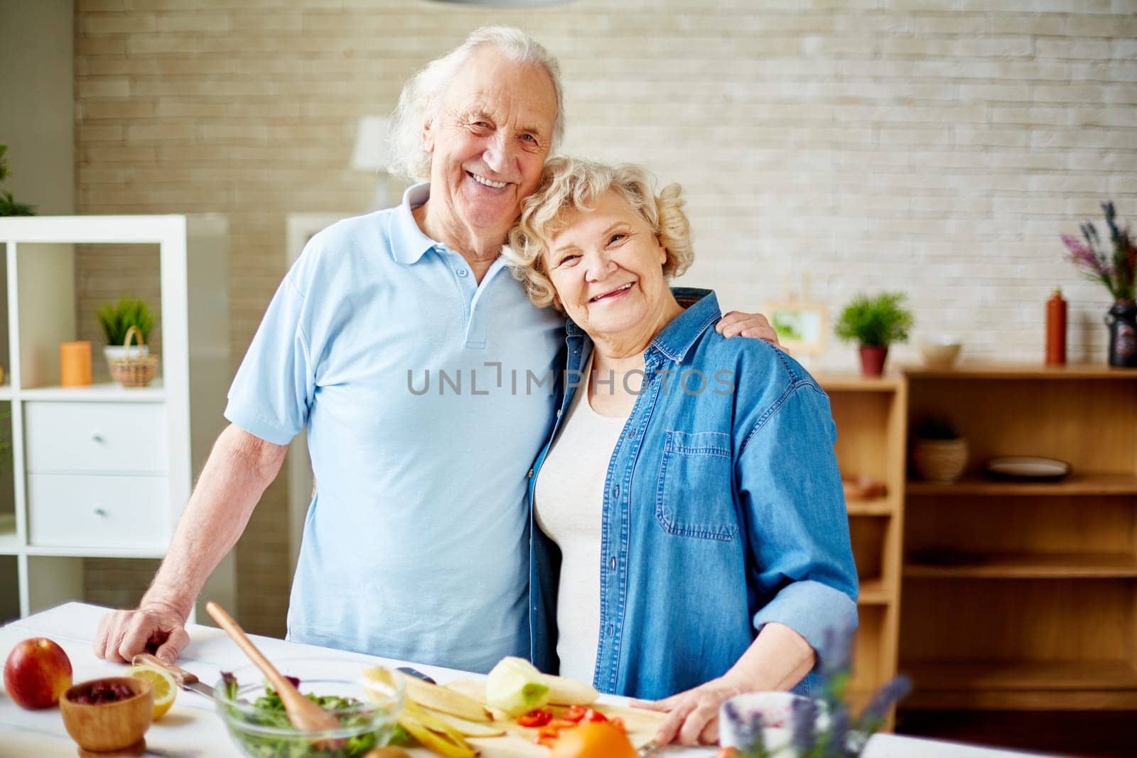 Cheerful seniors standing by table with fruits and looking at camera in the kitchen