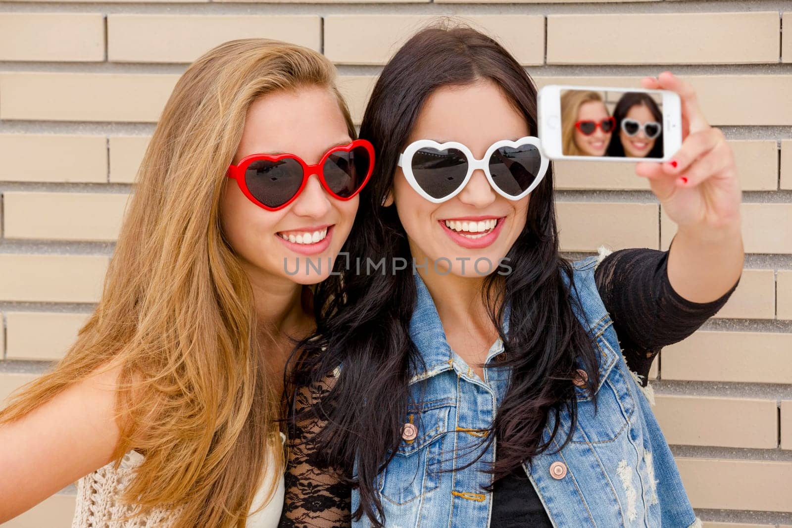 Two beautiful and young girlfriends taking pictures, in front of a brick wall