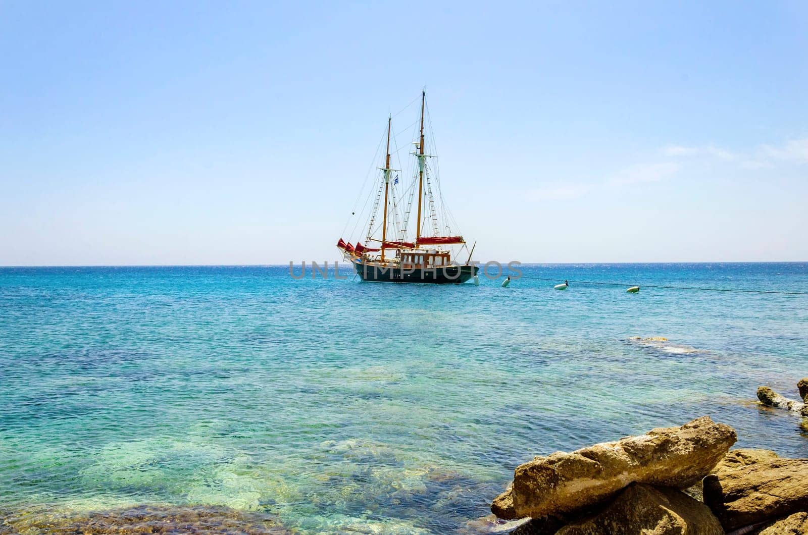 A traditional wooden sail boat anchored on the coast of Mykonos, Greece. The wind sails are down and it is tied on the rocks by a rope. A very typical greek island summer holiday scene on crystal clear blue sea.