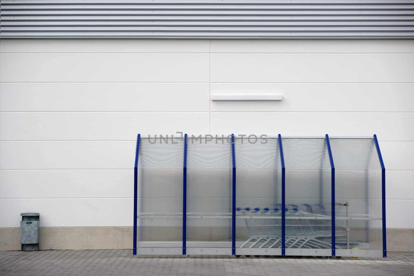 A plastic shelter with shopping baskets and a garbage can on the wall of a shopping center.