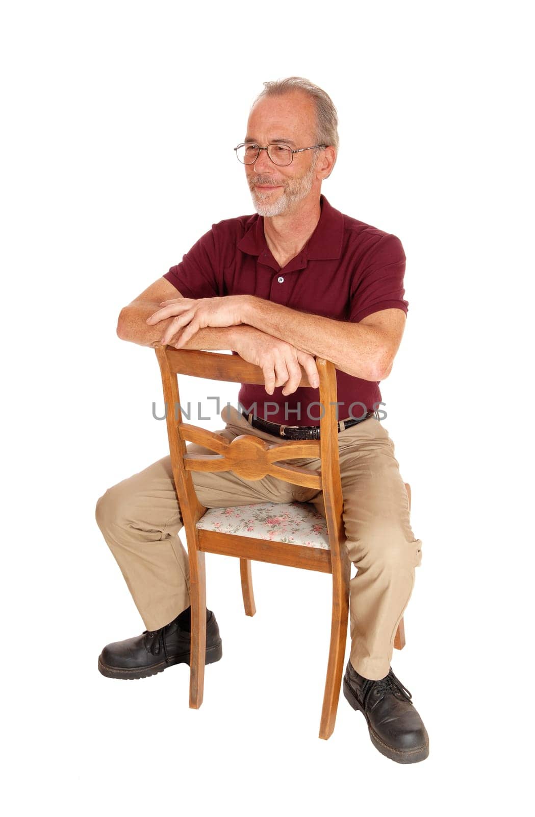 A serious older man in beige pants and burgundy t-shirt sitting backwards.on a chair, isolated for white background..