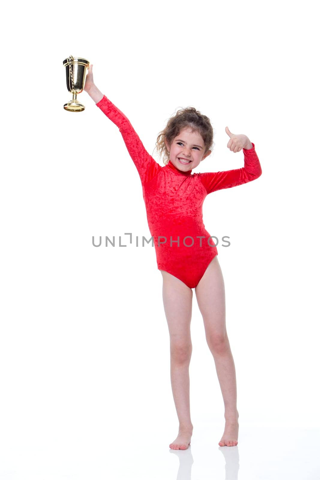 Young gymnast in a red leotard holding up a trophy on a white background. She is looking at the camera and smiling.