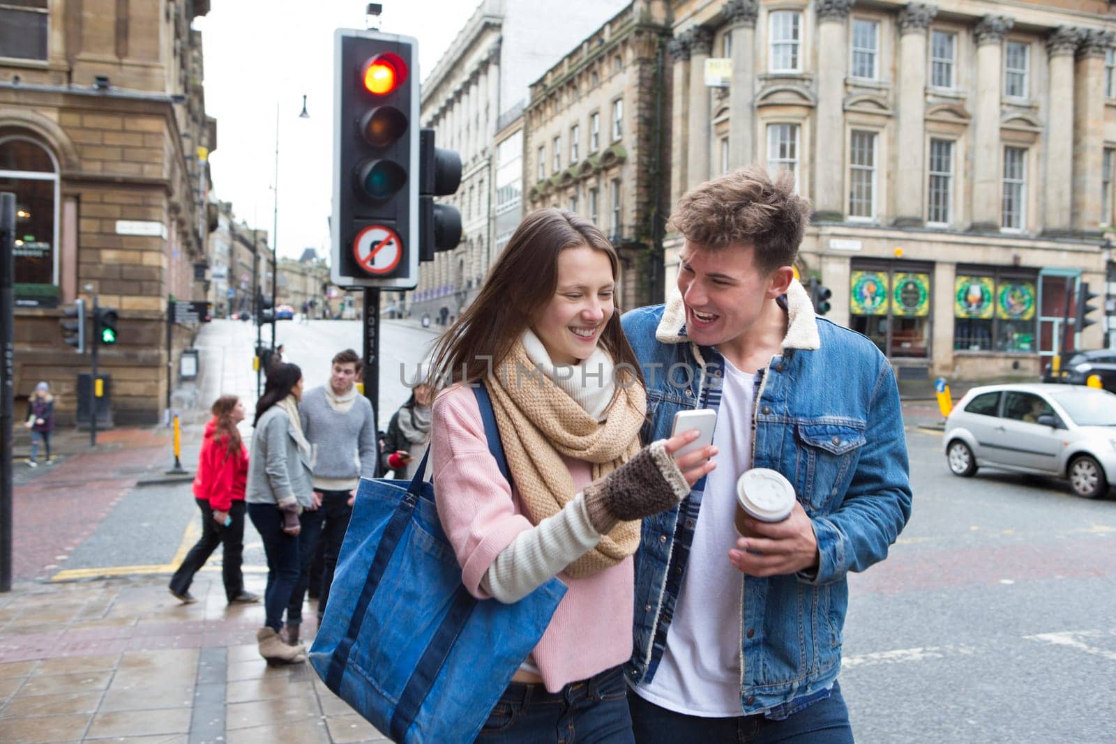 A young couple can be seen walking down a street looking at a smartphone together. Other young adults can be seen behind them.