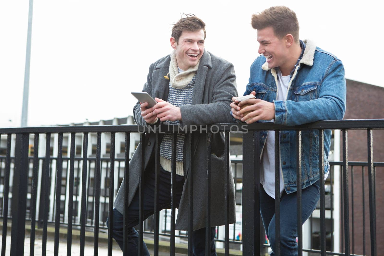 Two young men talking in the city. One is holding a smartphone, the other is holding a disposable coffee cup.