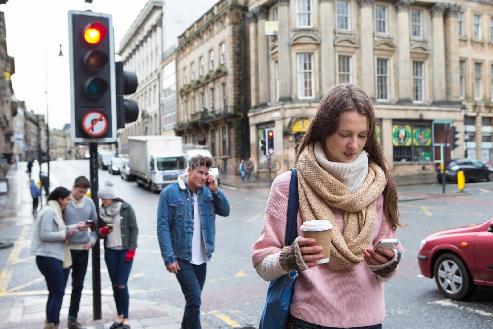 A young woman can be seen walking along a city street with a smart phone. Other young adults are in the background using smart phones.