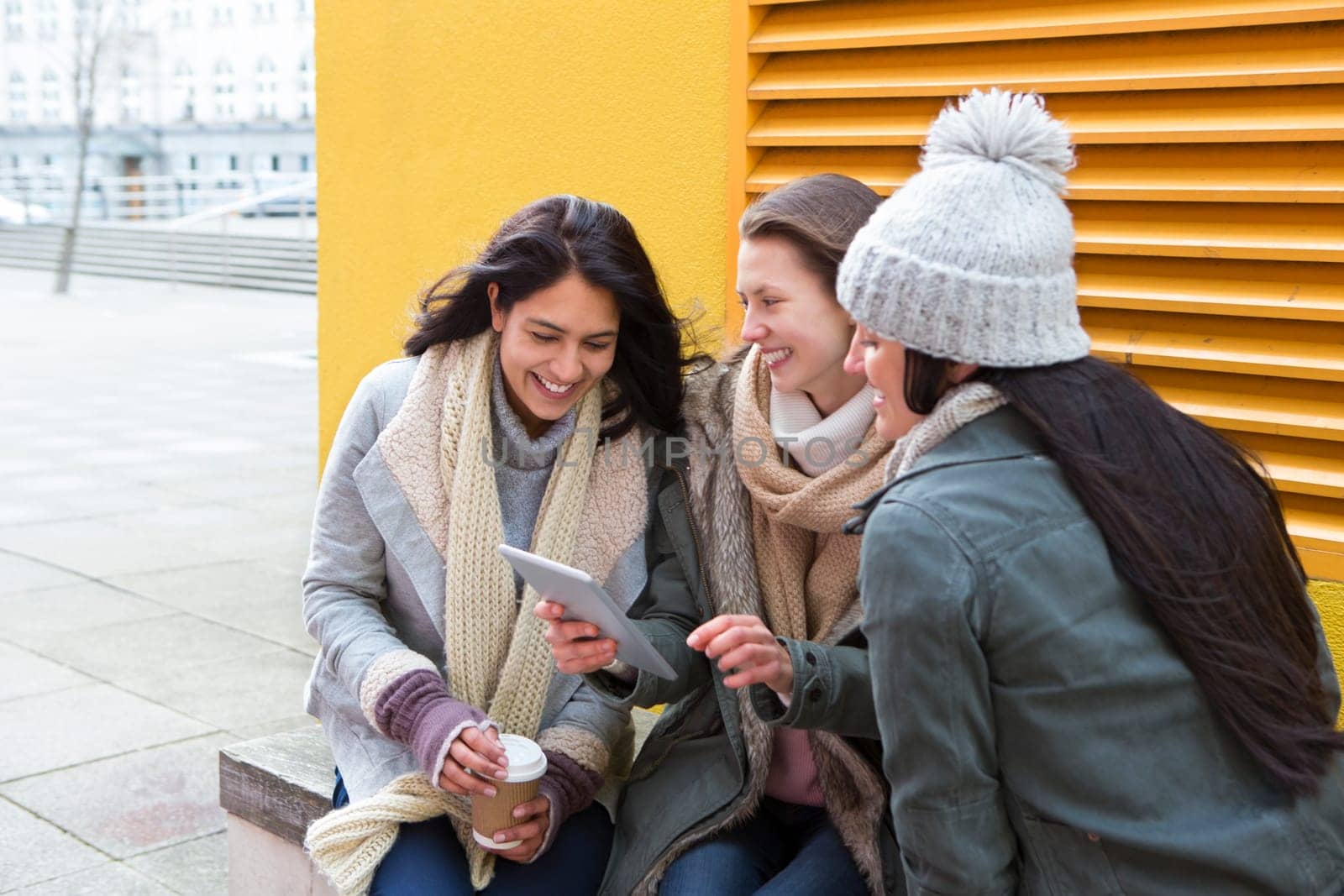 Three young women sitting in the city, looking at something on a digital tablet.