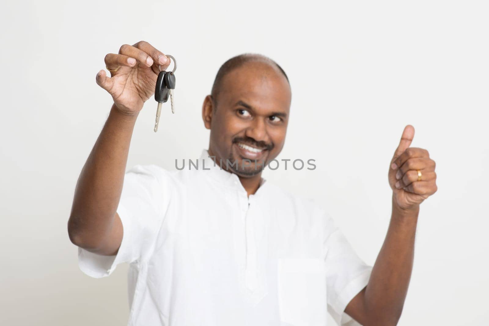 Portrait of Indian man holding new house key and thumb up, real estate property agent concept, focus on key, standing on plain background with shadow.
