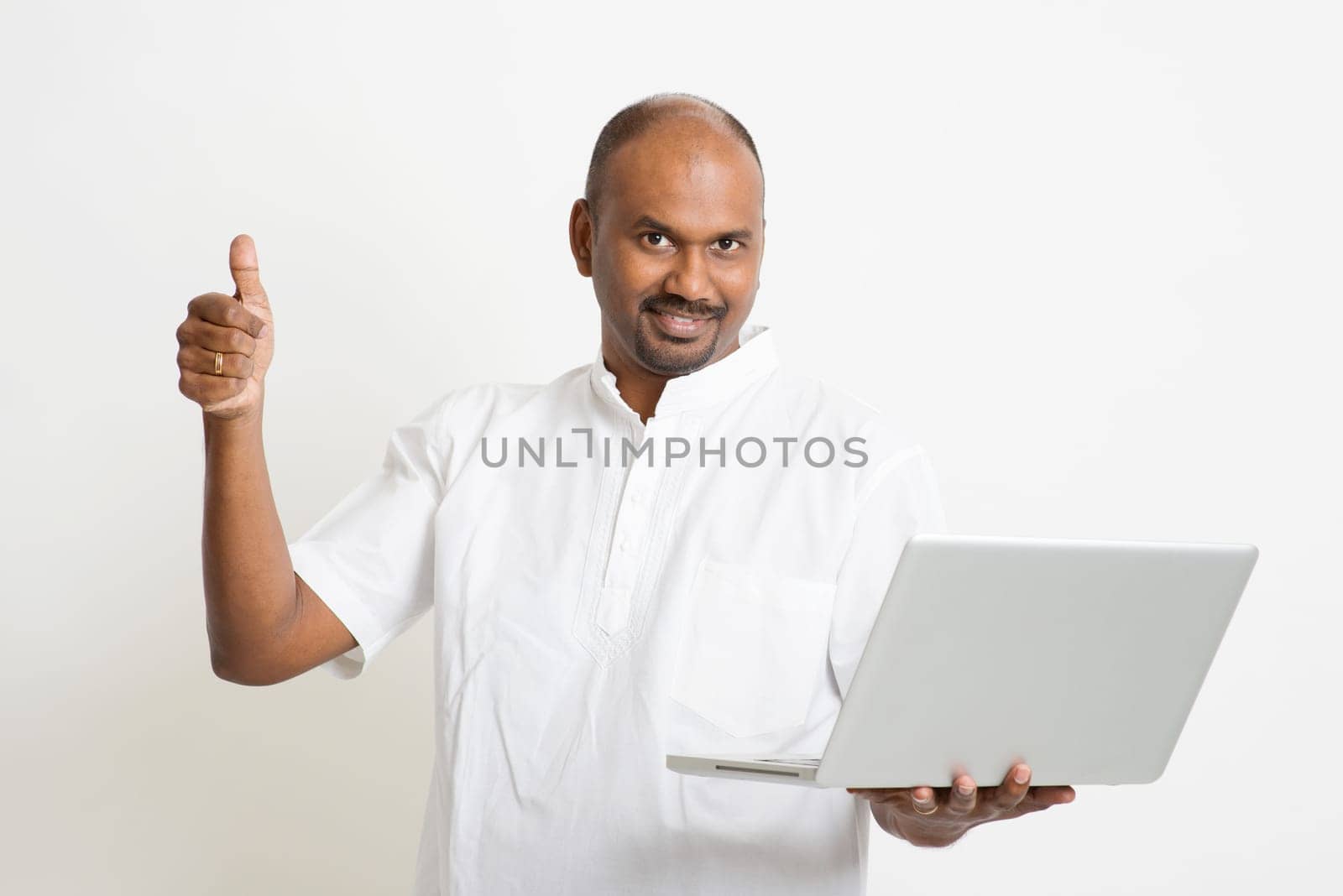 Portrait of mature casual business Indian man using laptop computer, looking at camera and thumb up, standing on plain background with shadow.