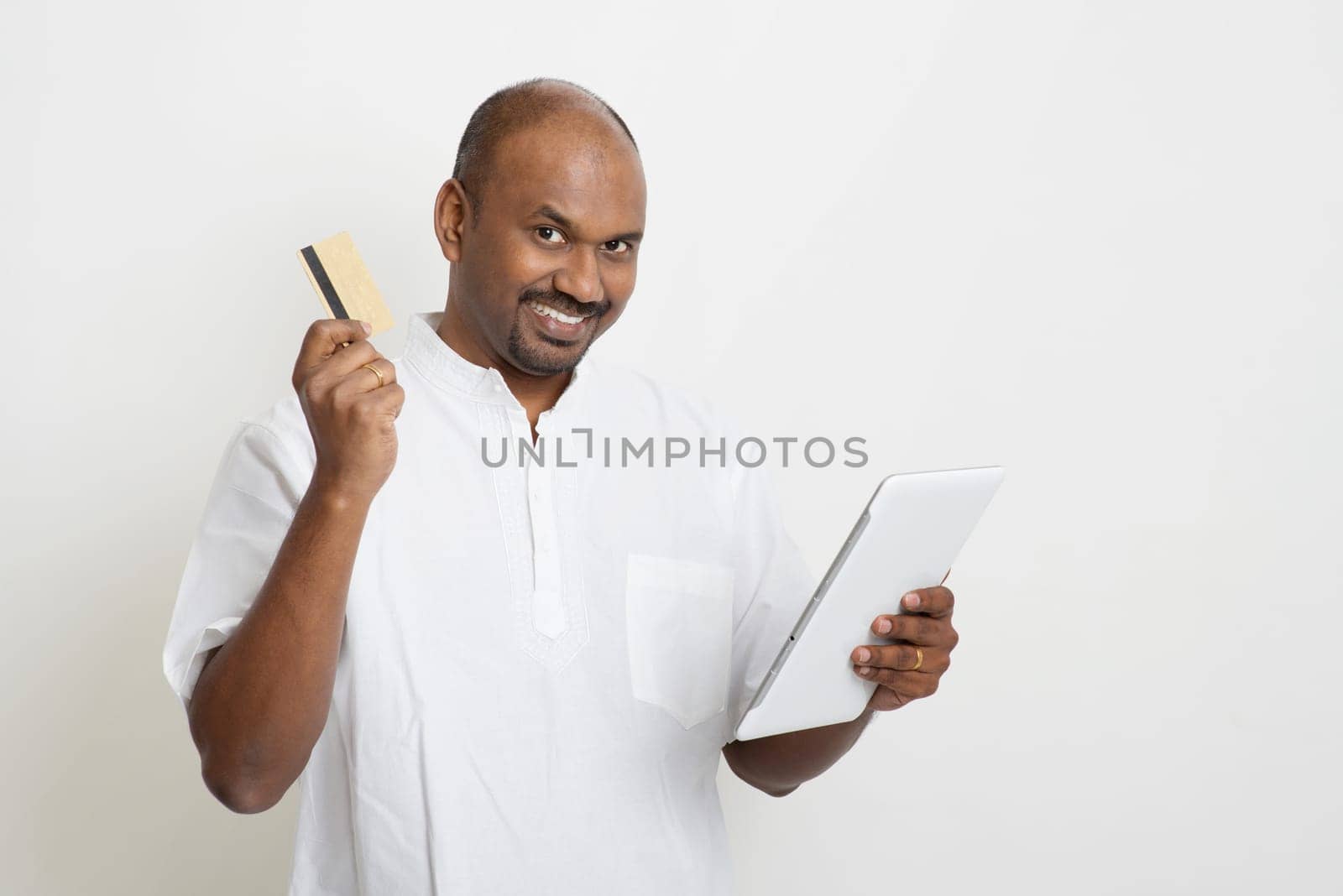 Portrait of mature Indian man shopping online using tablet pc, making payment by credit card. Asian man standing on plain background with shadow and copy space.