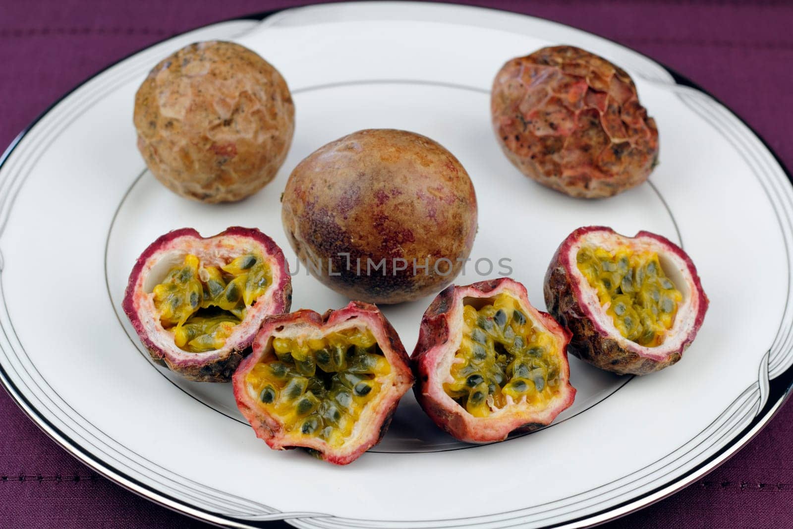 Three whole and four halves of a purple brown skinned passion fruit with yellow pulp  surrounding edible dark green seeds on a round white plate placed on a dark purple place mat.