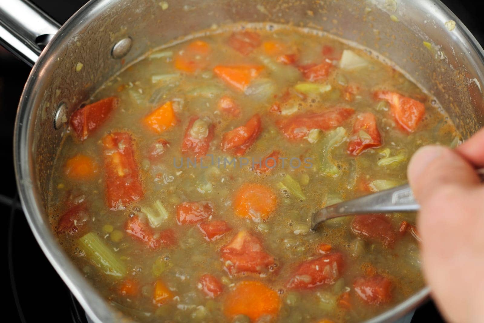 A hand of a caucasian man seen stirring lentil soup that has tomatoes, carrots and celery in the small stainless steel pan on a black glass stove top close up..