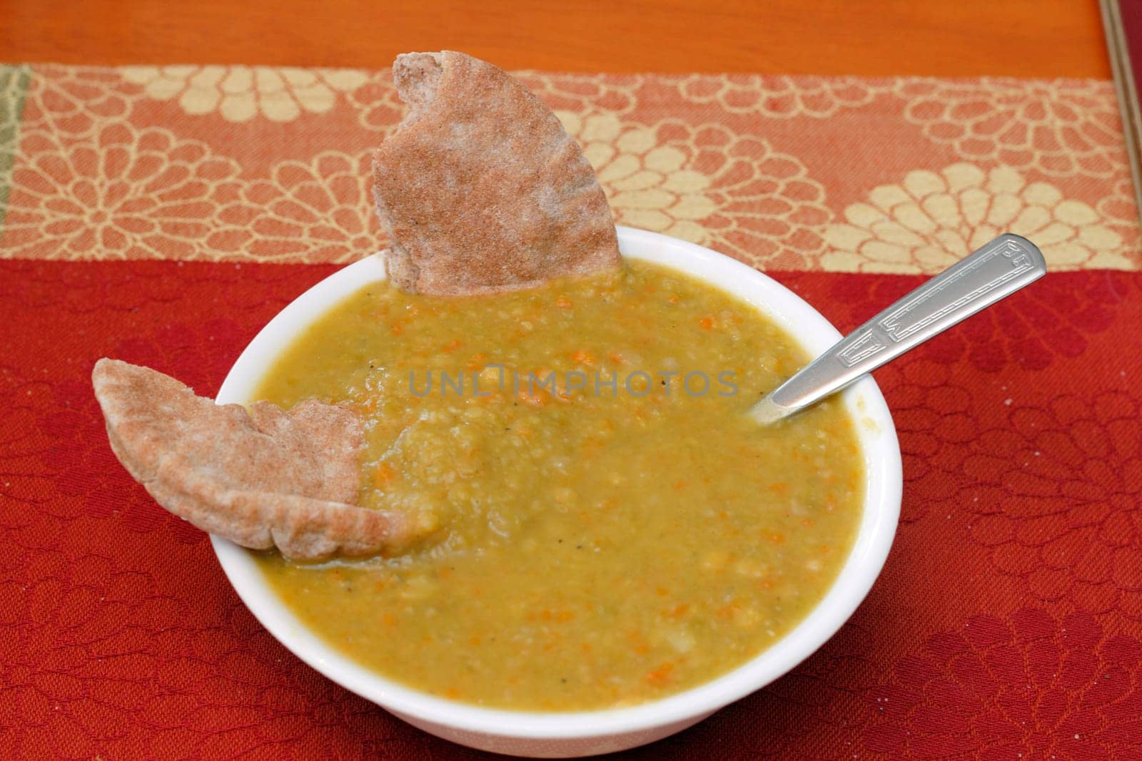 One bowl of split pea soup made with carrots. Two pieces of whole wheat pita bread and a spoon are dipped in split pea soup on a colorful place mat on a wooden table.