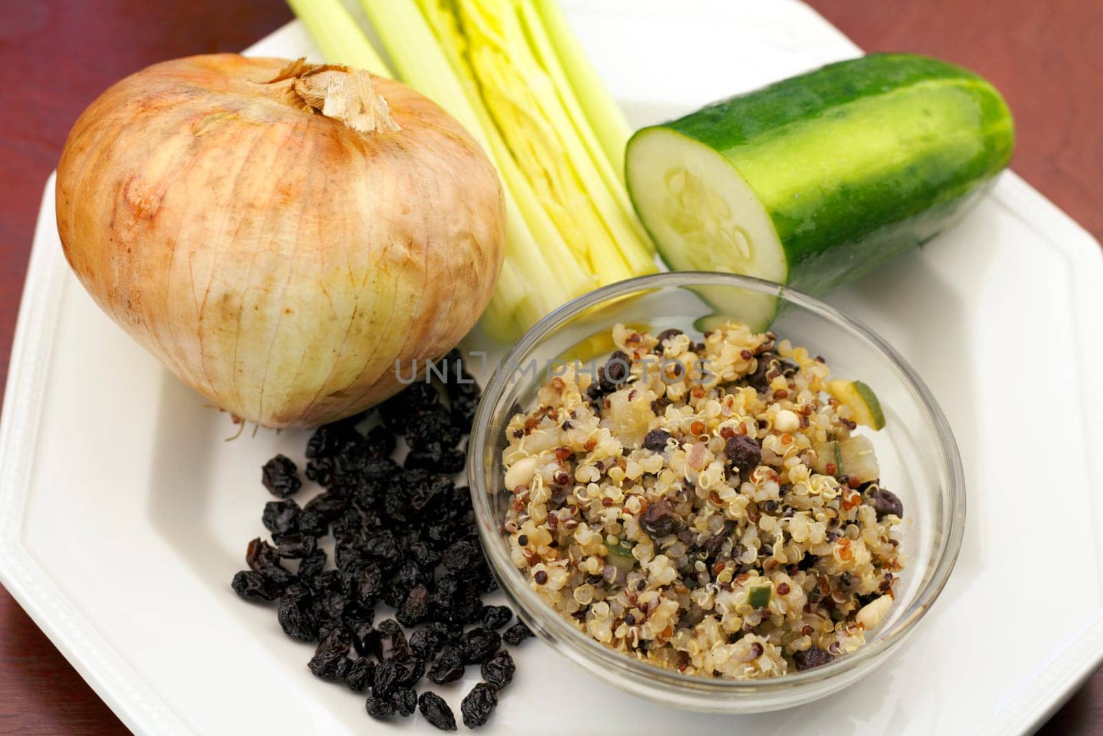 In a small round bowl is a cooked tricolor quinoa recipe surrounded by a bunch of black currants, a yellow onion, celery stalks and a half a cucumber on a plate resting on a wood surface..