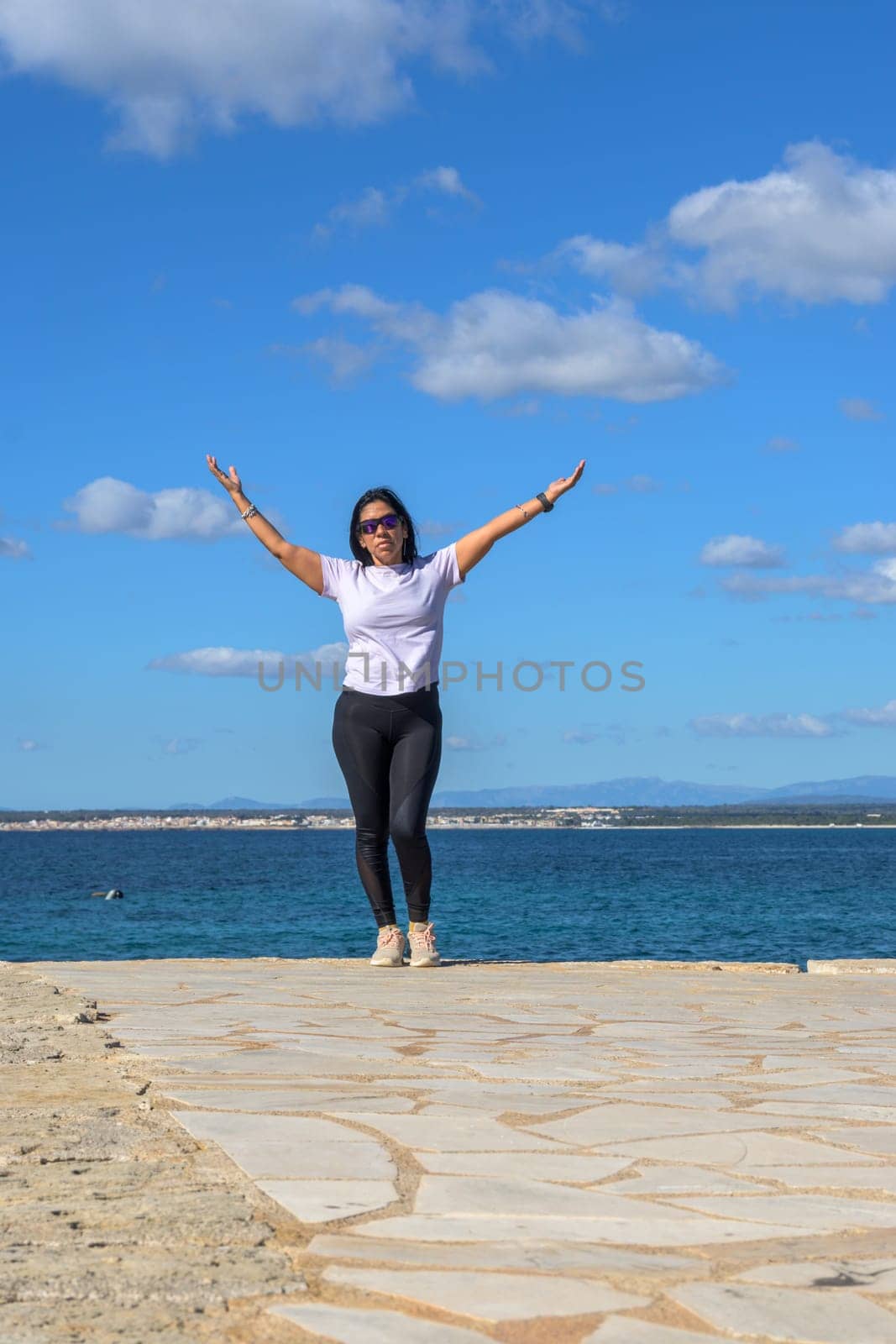 A woman stands on a rocky edge by the sea, arms open wide under a blue sky with scattered clouds, beautiful latina woman by the sea in Colonia San Jordi, Balearic Islands, Majorca, Spain,