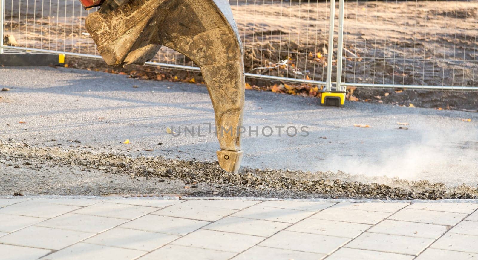 Excavator Ripping Asphalt with fence behind it.