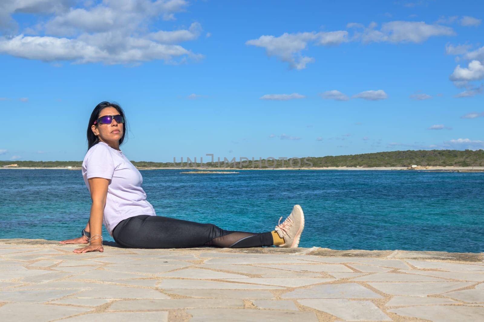 Woman reclining by the ocean, in a relaxed posture, wearing sunglasses under the sunny sky, beautiful latina woman by the sea in Colonia San Jordi, Balearic Islands, Majorca, Spain