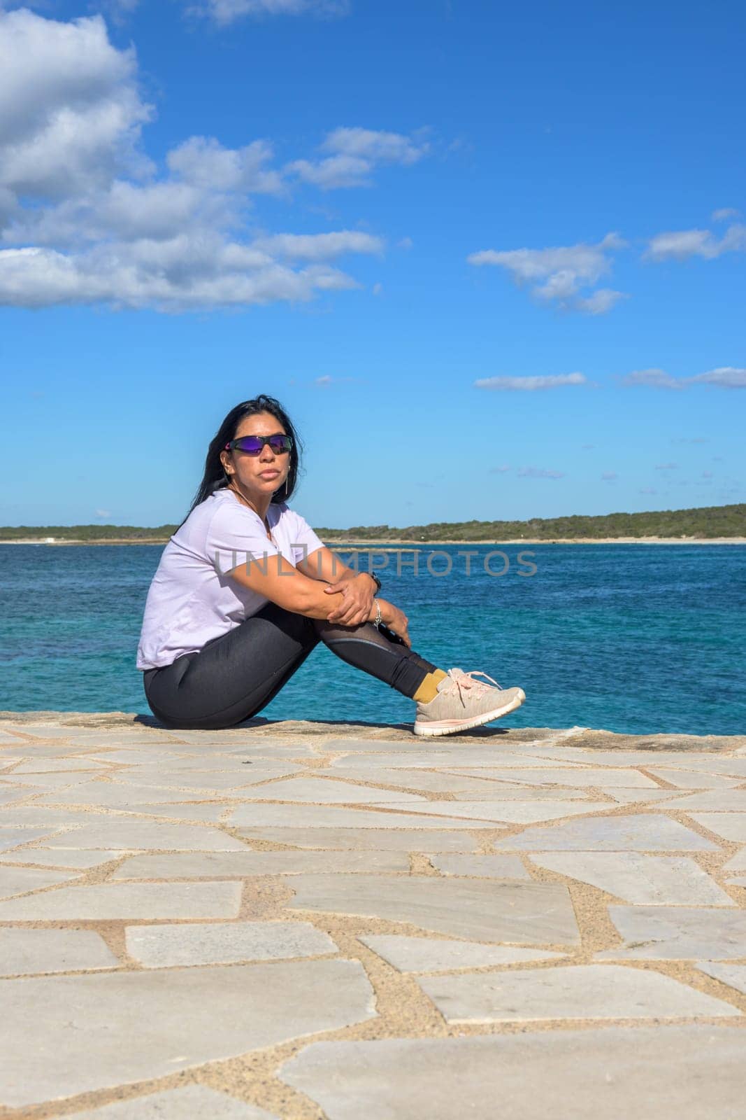 Latin woman wearing sunglasses and sportswear sitting by the sea in Colonia de Sant Jordi, Mallorca, during a sunny spring day, enjoying the scenery.