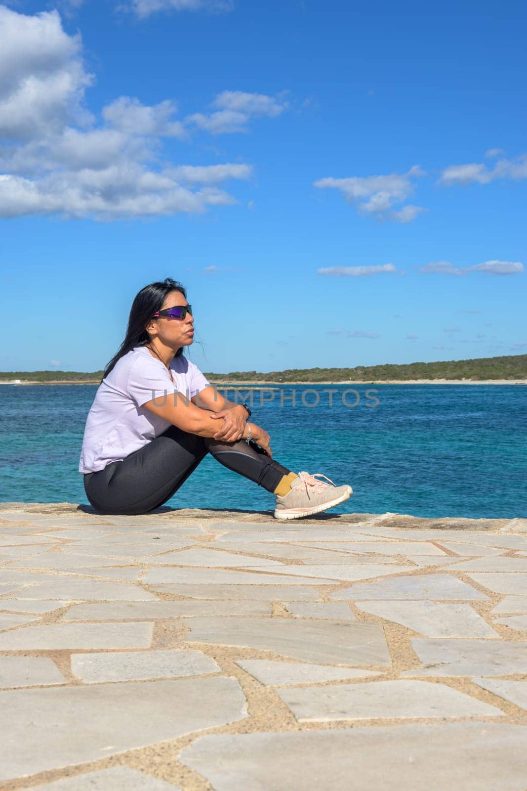 Latina woman, 48 years old, sitting on a pier in Colonia de Sant Jordi, Mallorca, Spain. A sunny early spring day by the turquoise sea, enjoying the peaceful coastal view.