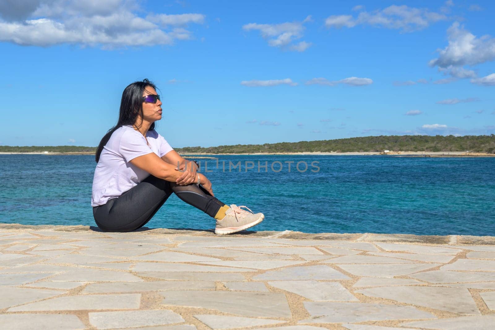 Latina woman, 48 years old, sitting on a pier in Colonia de Sant Jordi, Mallorca, Spain. A sunny early spring day by the turquoise sea, enjoying the peaceful coastal view.