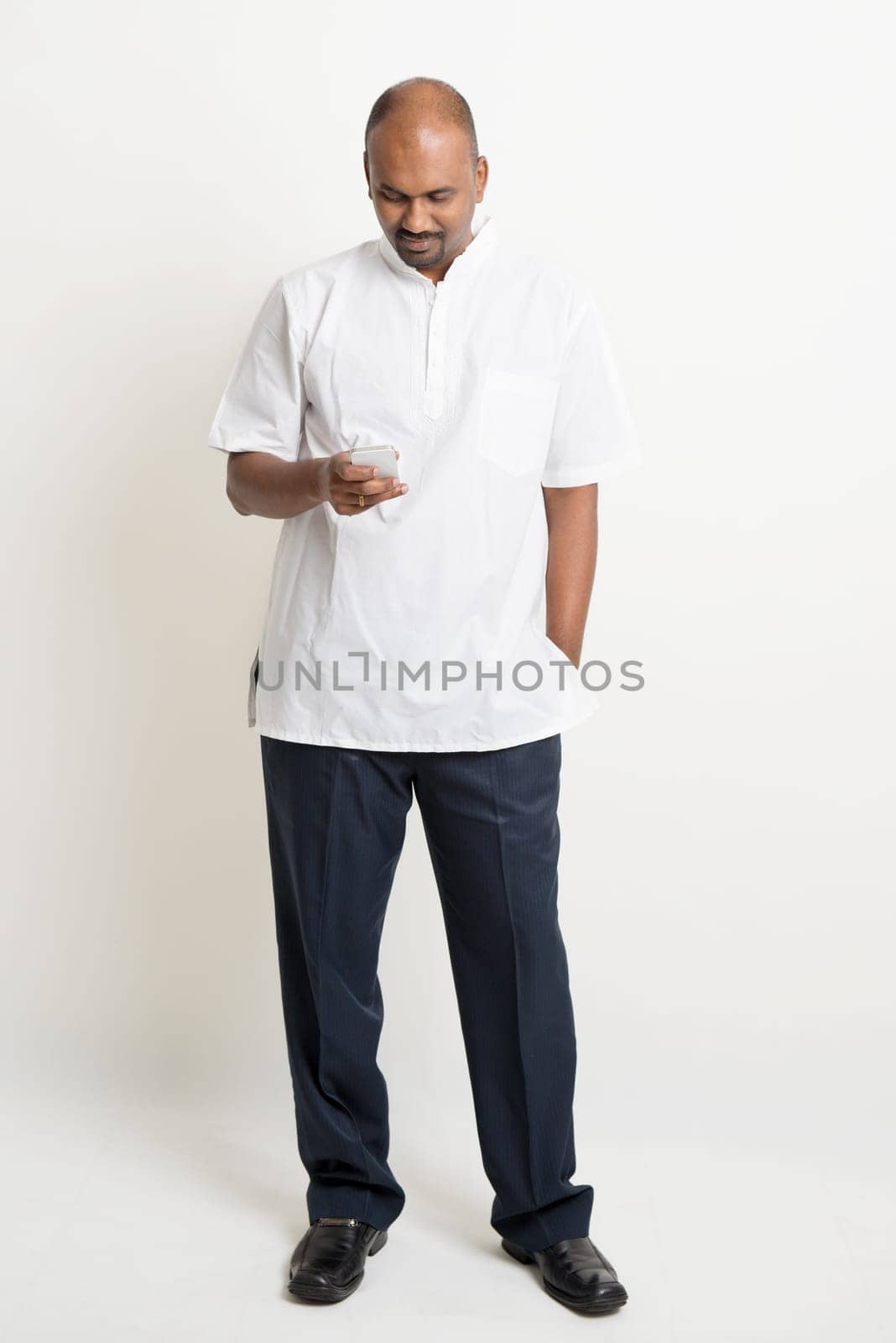 Portrait of full length mature casual business Indian man using smartphone, mobile apps concept, standing on plain background with shadow.