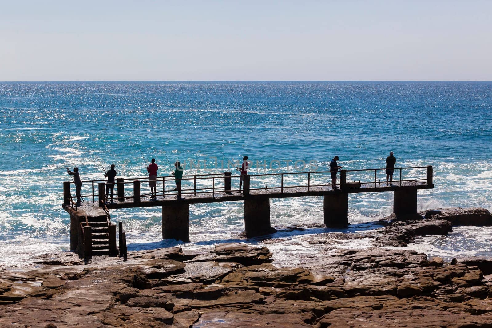 Fishermen on concrete pier jetty with blue ocean water along rocky coastline landscape.