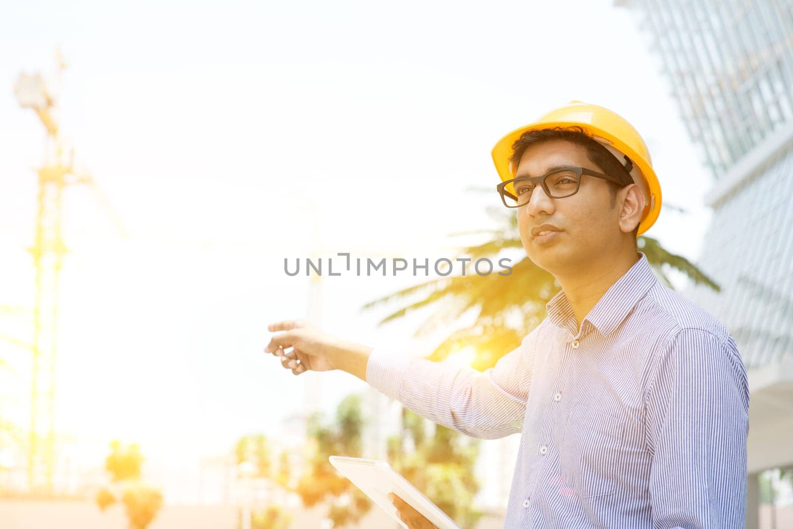 Portrait of a smiling Indian male contractor engineer with hard hat pointing to a crane at construction site