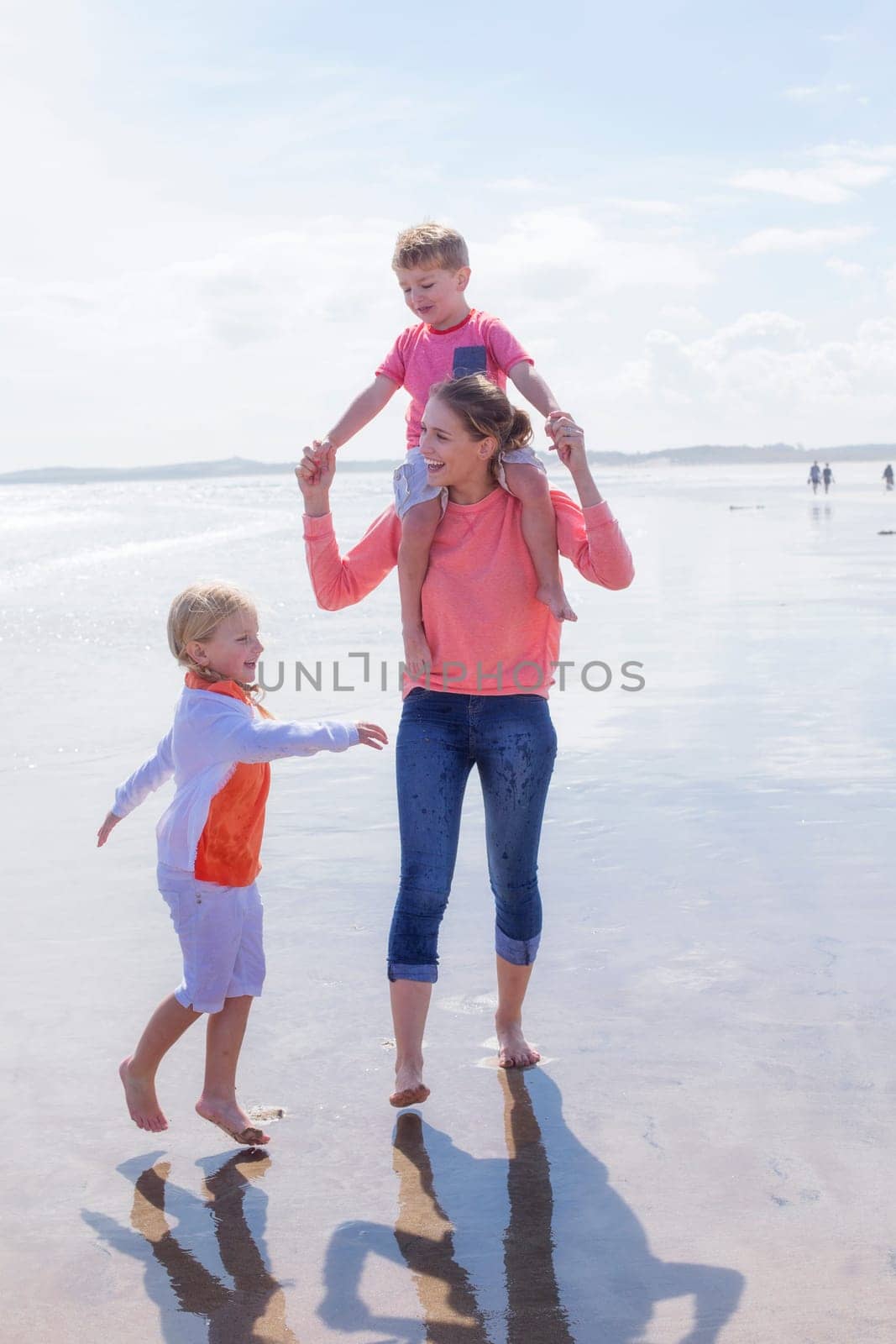 Happy single parent family walking along the beach. They are all laughing and smiling and walking across the waters edge. The daughter is walking alongside her mother while her son is on her shoulders.