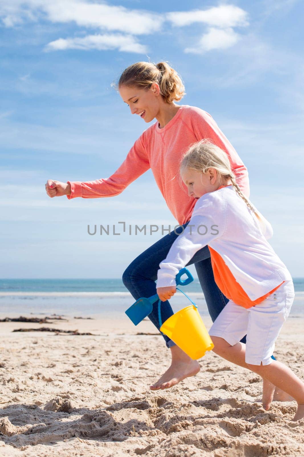 Young mother and daughter running along the beach with a bucket and spade. They are both laughing and smiling.