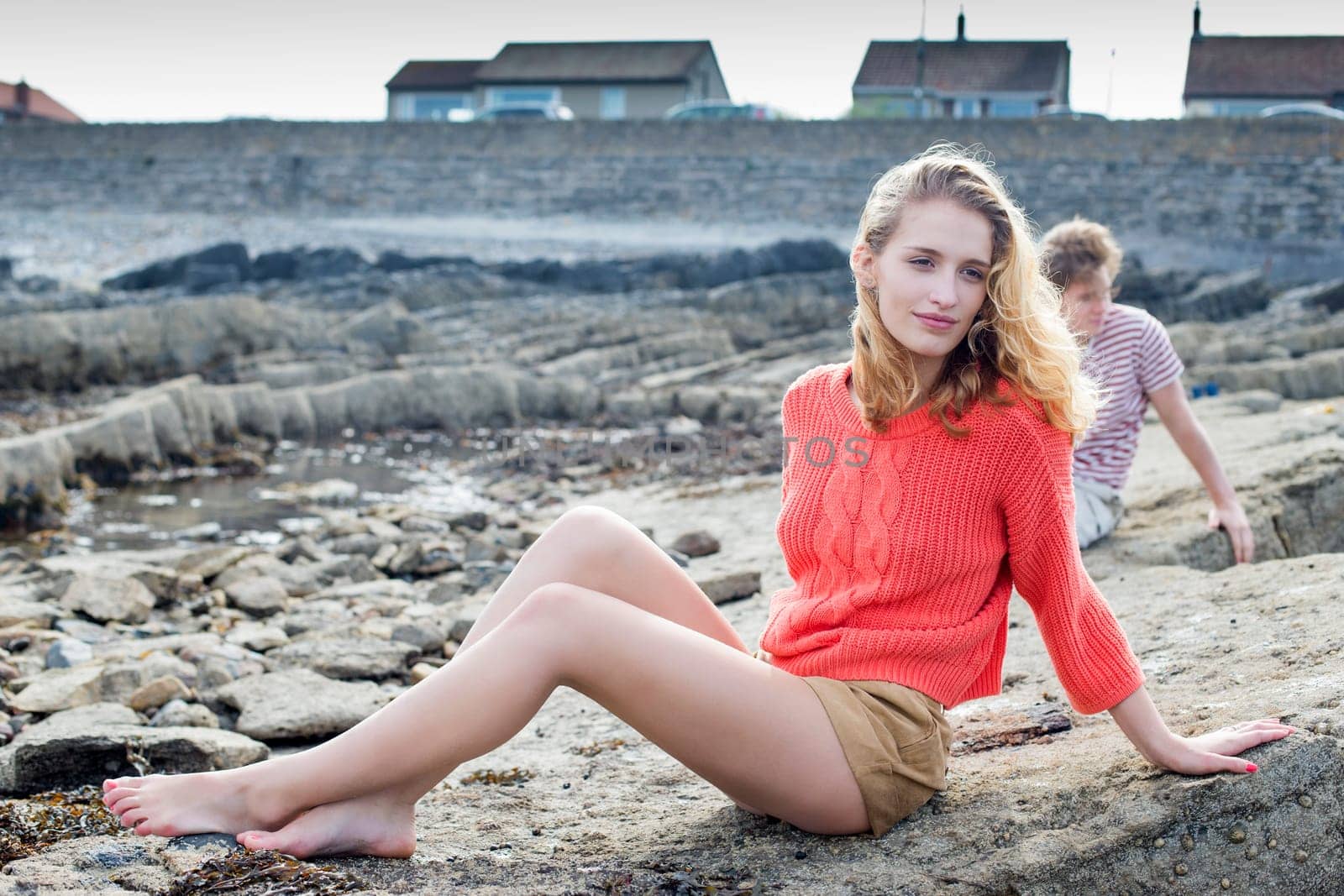 Young girl sitting on a rock at the beach wearing casual clothing. Her boyfriend can be seen in the background.