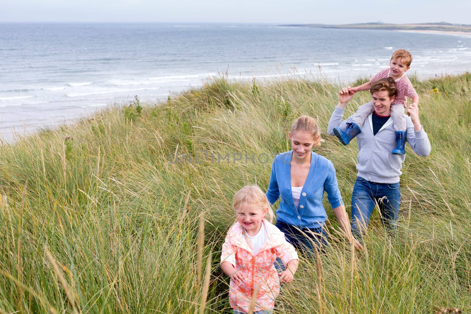 A young family of four walking through long grass next to the beach. They are wearing casual clothing and are smiling.