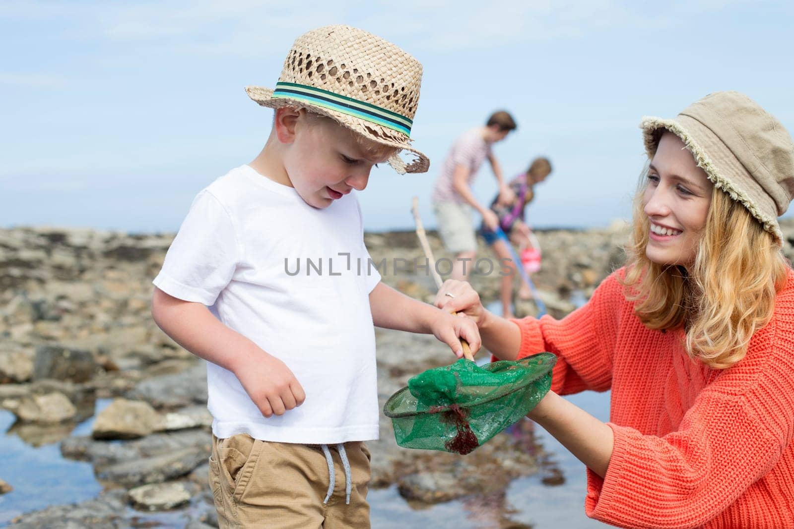 Young family at the beach searching the rock pools. The mother and her young son are in the foreground and she is holding the crab in the net to show her son.