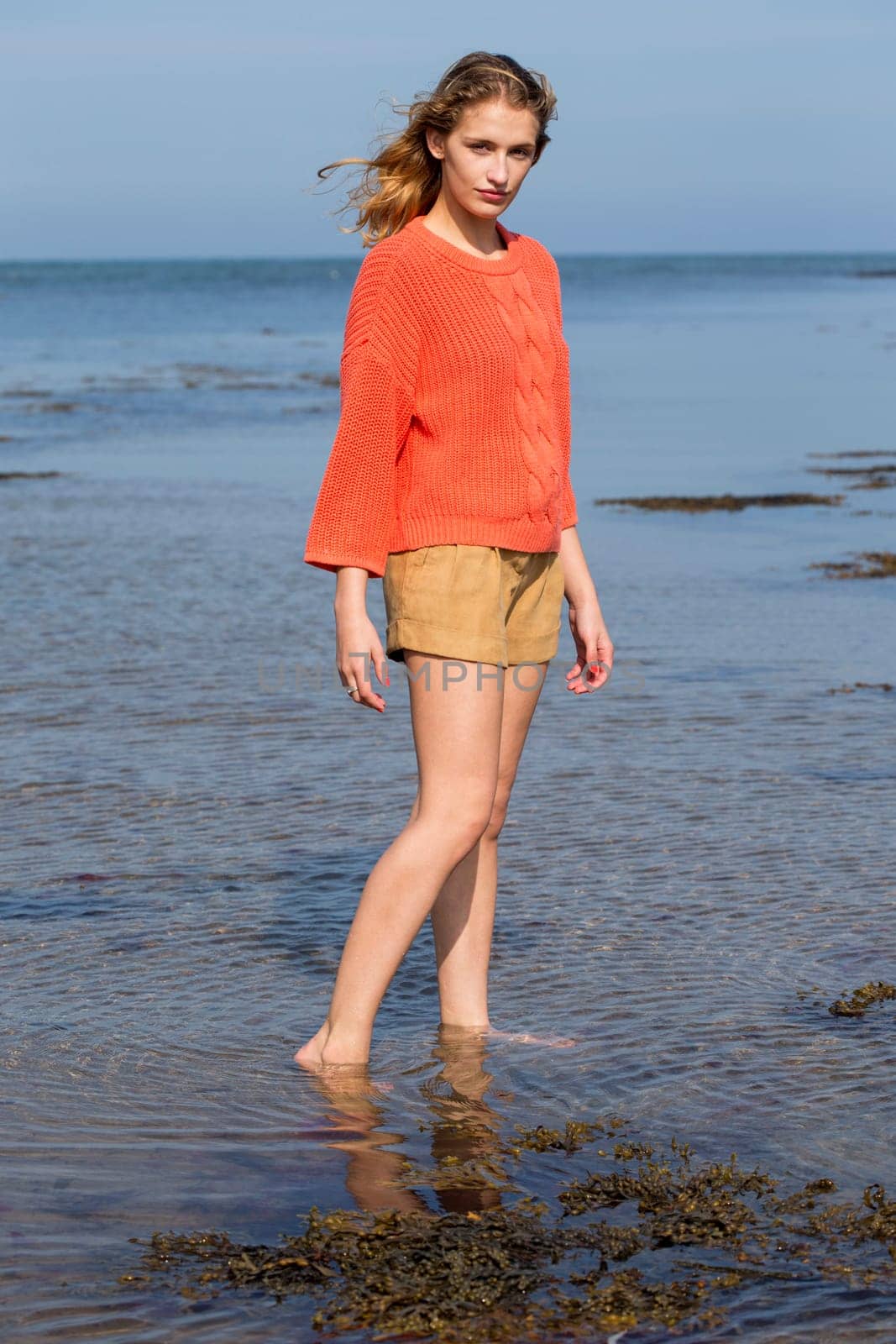 Portrait of a beautiful young woman at the beach. She is looking at the camera and wearing casual clothing while standing in the sea.