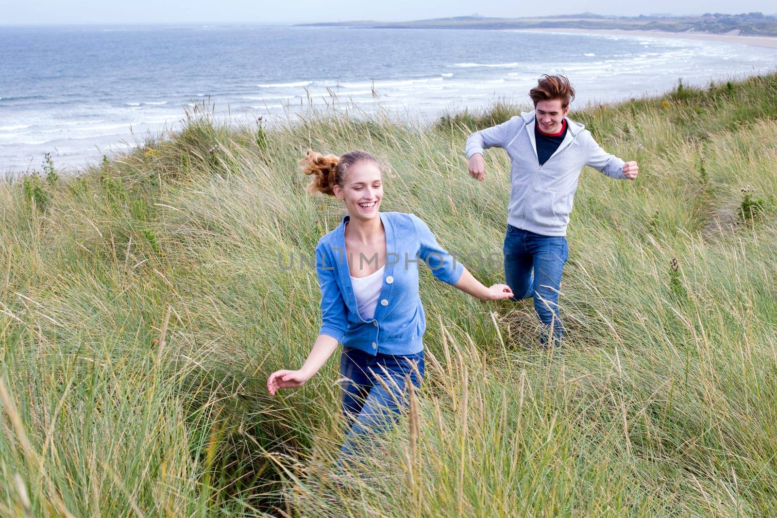 Young couple running through long grass with the background of the beach. They are both smiling and wearing casual clothing.