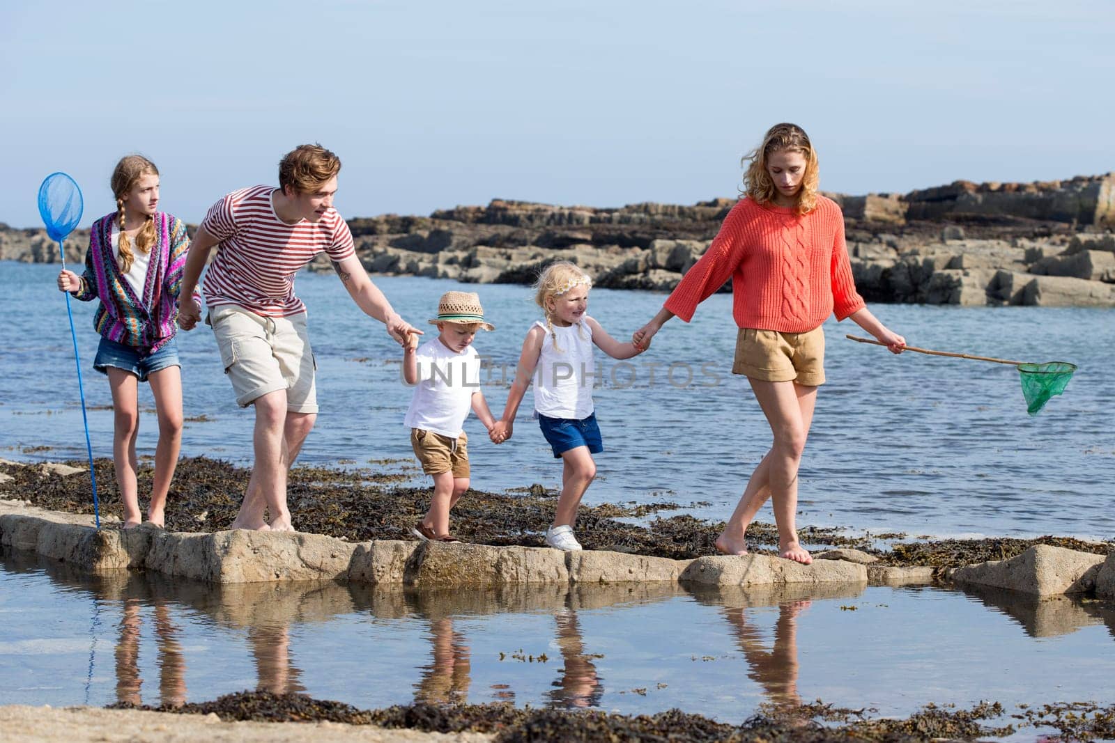 Young family walking along the rocks at the beach. They are all wearing casual clothing and holding hands and nets.